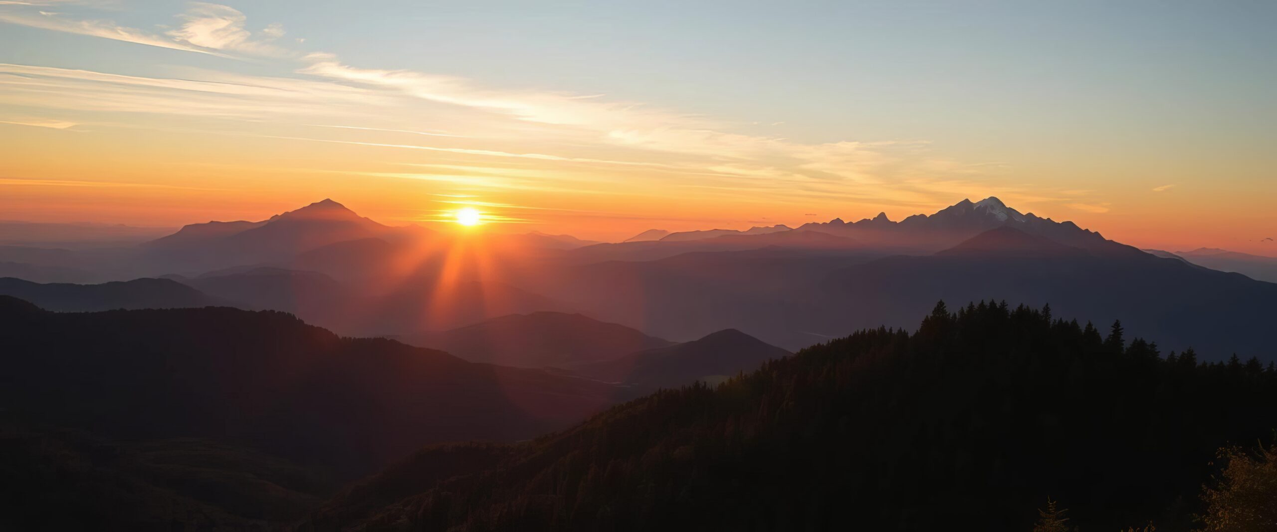 „Weitläufige Berglandschaft bei Sonnenaufgang: Die Sonne erscheint am Horizont zwischen zwei Bergmassiven und taucht Himmel und Täler in warmes orangefarbenes und rosiges Licht. Silhouetten mehrerer Bergketten liegen gestaffelt im Vorder- und Hintergrund, während feine Wolkenstreifen den farbigen Himmel durchziehen.“